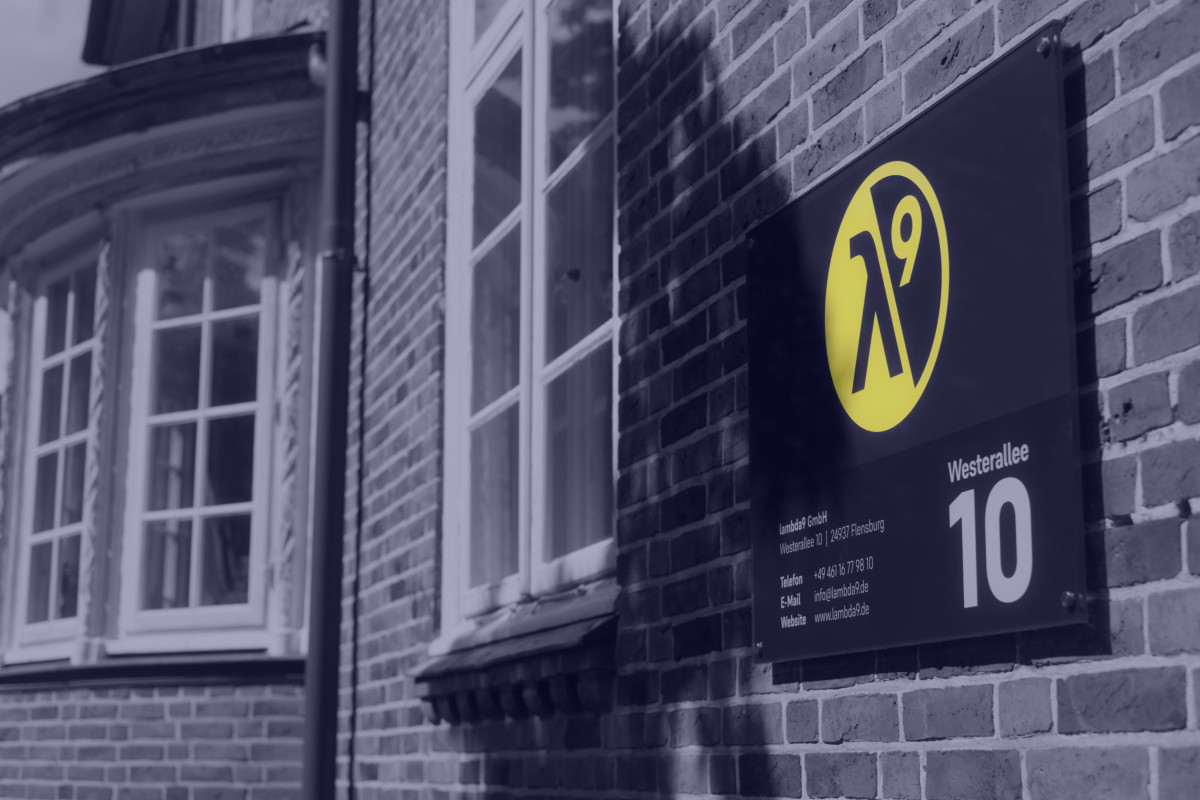 Vor einer Häuserwand ist ein Schild mit Logo und Adresse der Firma lambda9 montiert.